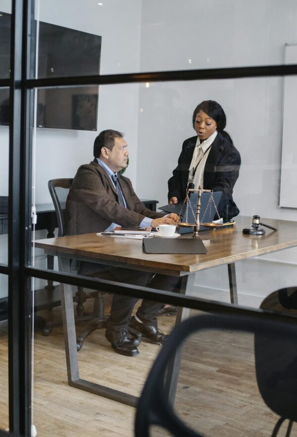 View through glass of serious black woman in formal suit explaining opinion to interested mature colleague in light office with judge gavel and scales on table